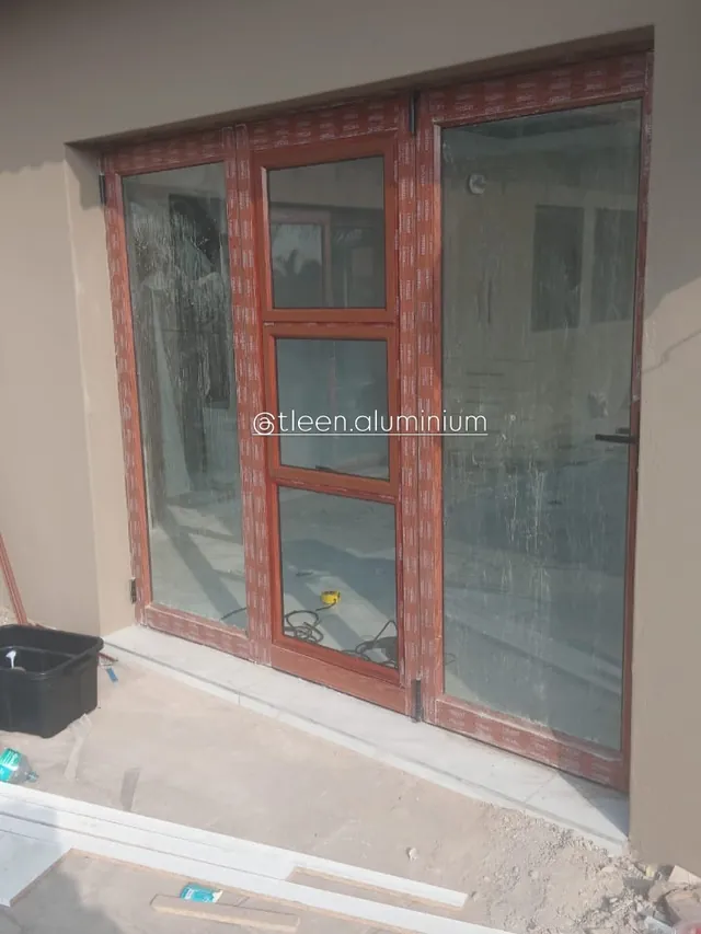 Wood-look aluminium door and two fixed side panes glazed into a finished opening, protective film still taped to every frame, floor screed not yet finished