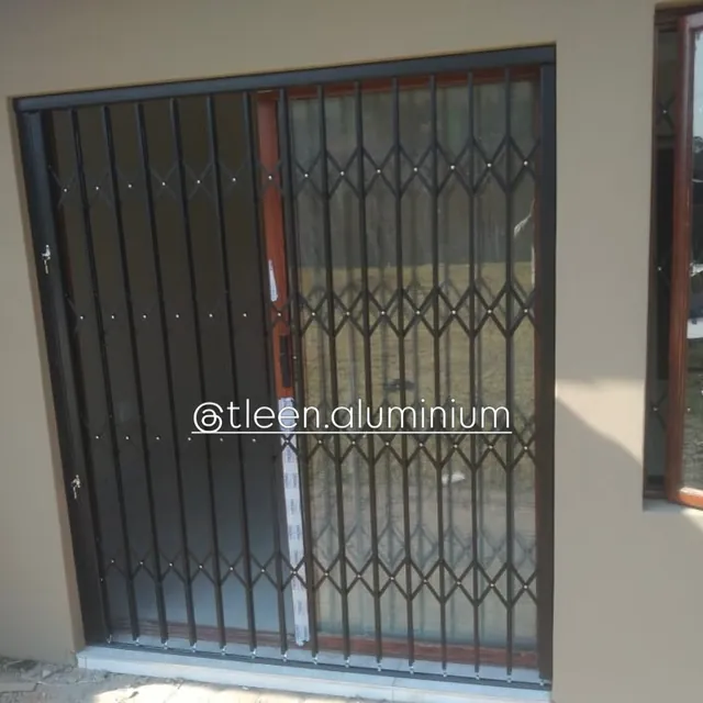 Charcoal trellis security gate over a wood-look aluminium door, protective film still on the frame, plaster made good around the opening